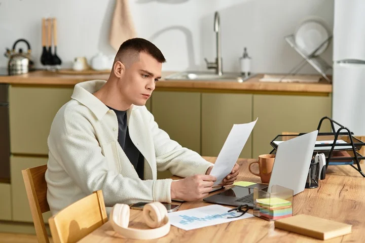 Young man reading a document at his kitchen table with laptop and phone nearby, arranging a medical certificate for Centrelink