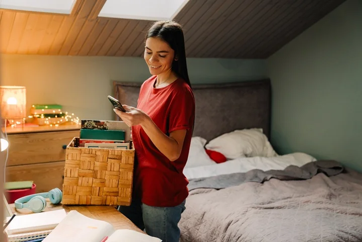 Student smiling while checking her phone next to a box of books in her bedroom, arranging a certificate for course deferral