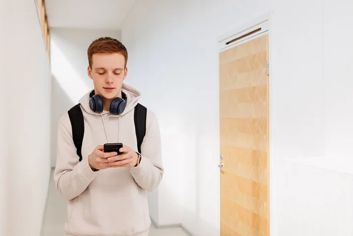Teenage student checking his phone in a bright school hallway with backpack on, arranging a medical certificate for school absence