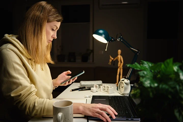 University student checking her phone at a lamp-lit desk with laptop open, arranging a medical certificate for special consideration