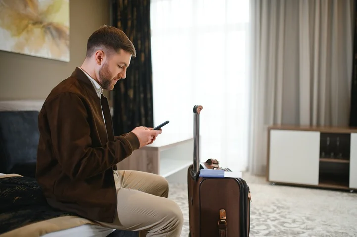 Man checking his phone while seated near his suitcase in a naturally lit hotel bedroom, preparing for his fit to fly certificate