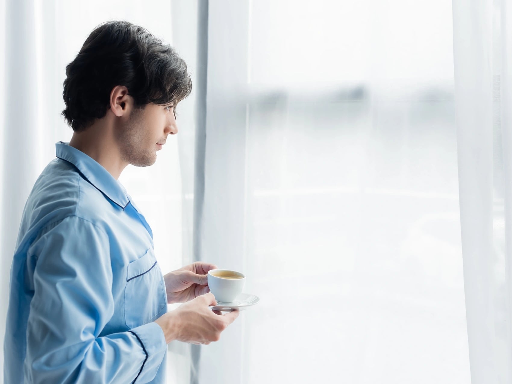 Man in blue pyjamas holding a cup of tea by a bright window early in the morning while home sick for the day