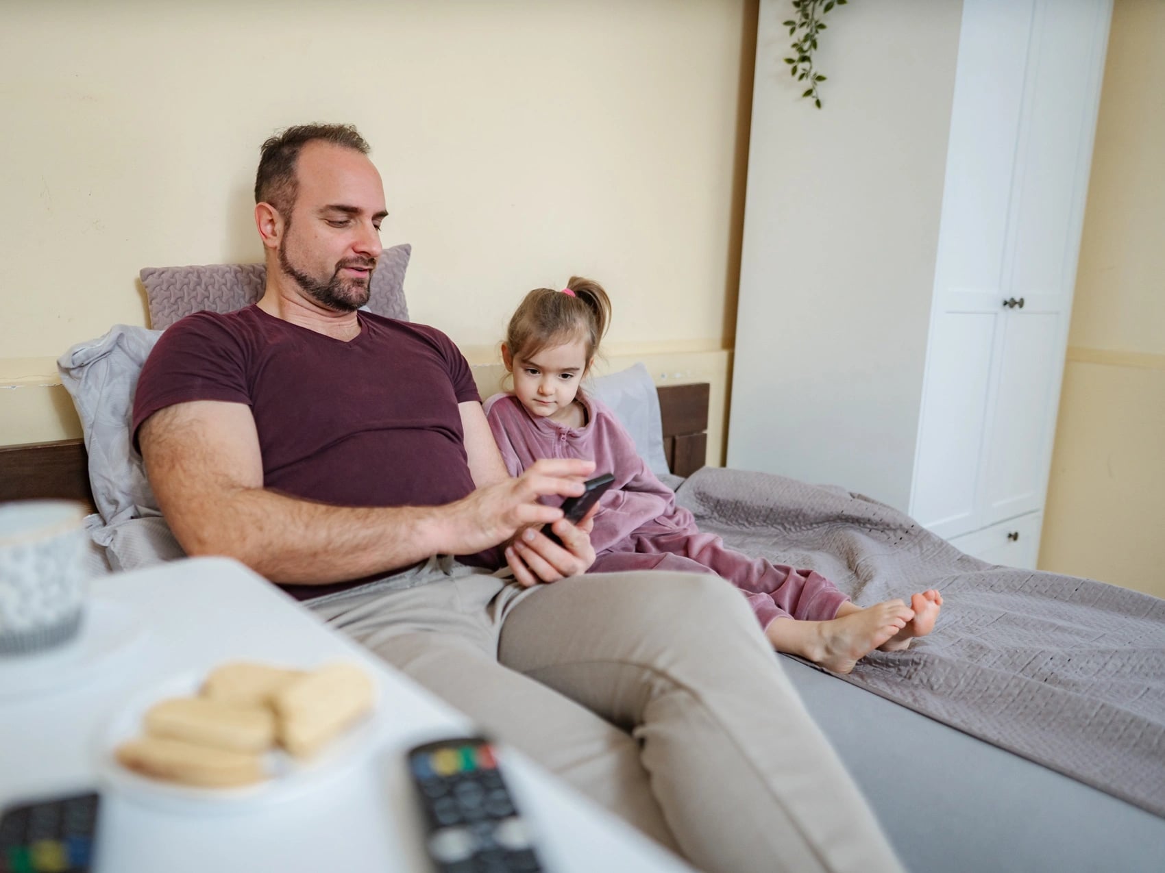 Father lying beside his young daughter as she rests in bed at home, holding his phone while caring for her