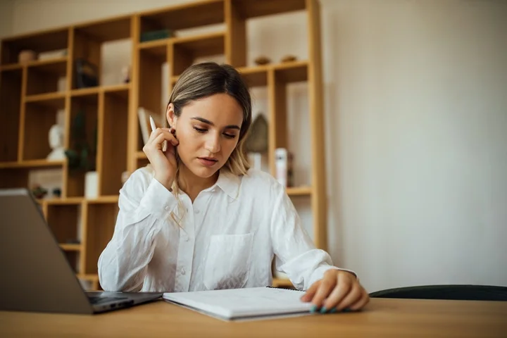 Woman reviewing notes at a bright home desk with her laptop open, preparing her carer's leave certificate for her employer