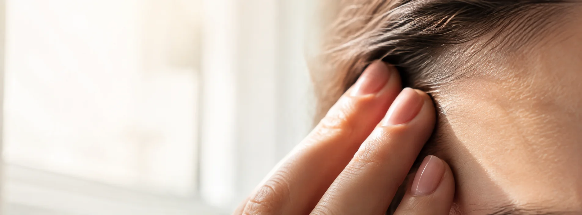 Close-up of fingers gently pressed to the temple with a softly blurred bright window behind, representing light sensitivity after concussion
