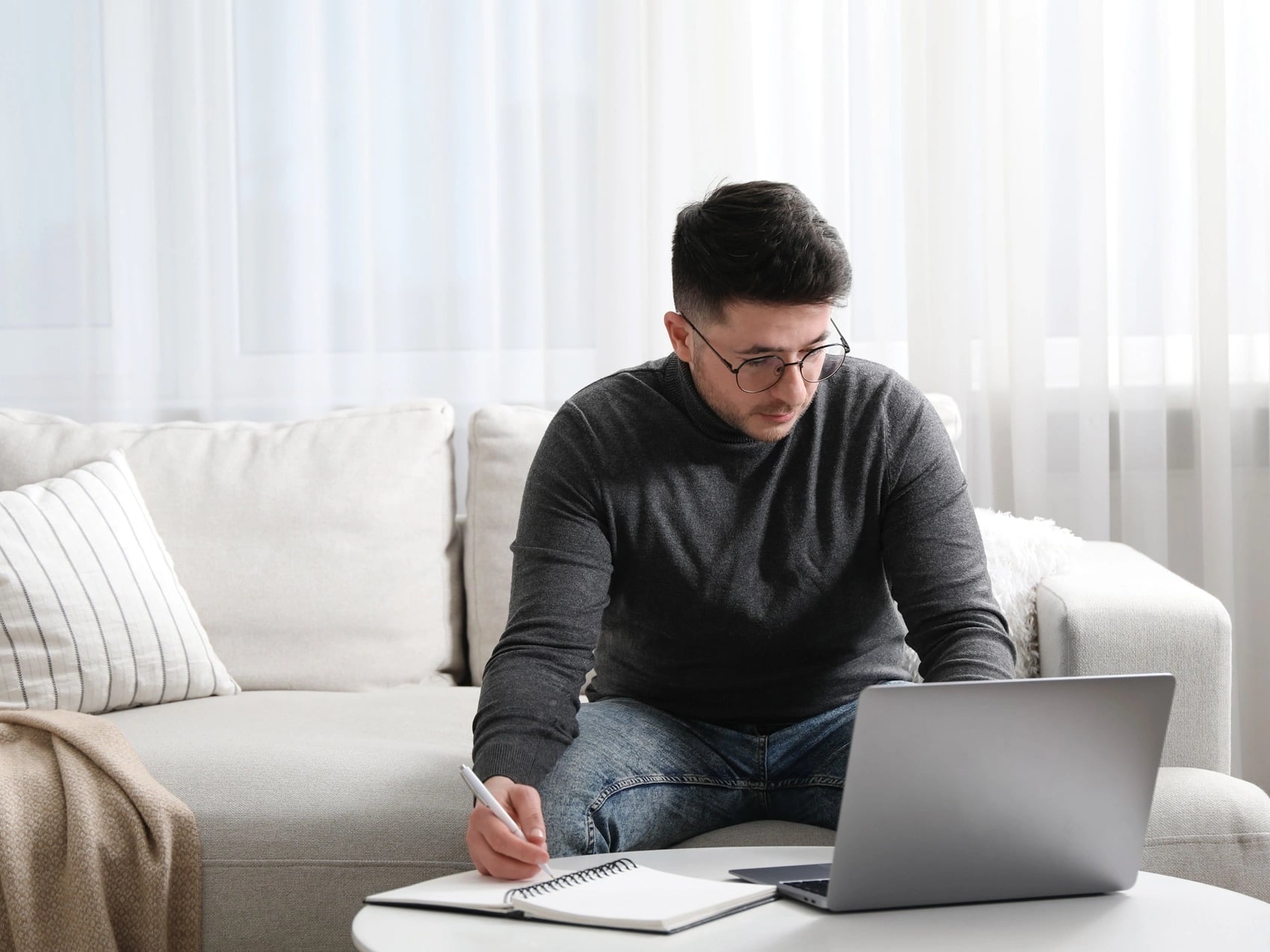 Student sitting on a sofa at home, writing notes beside an open laptop