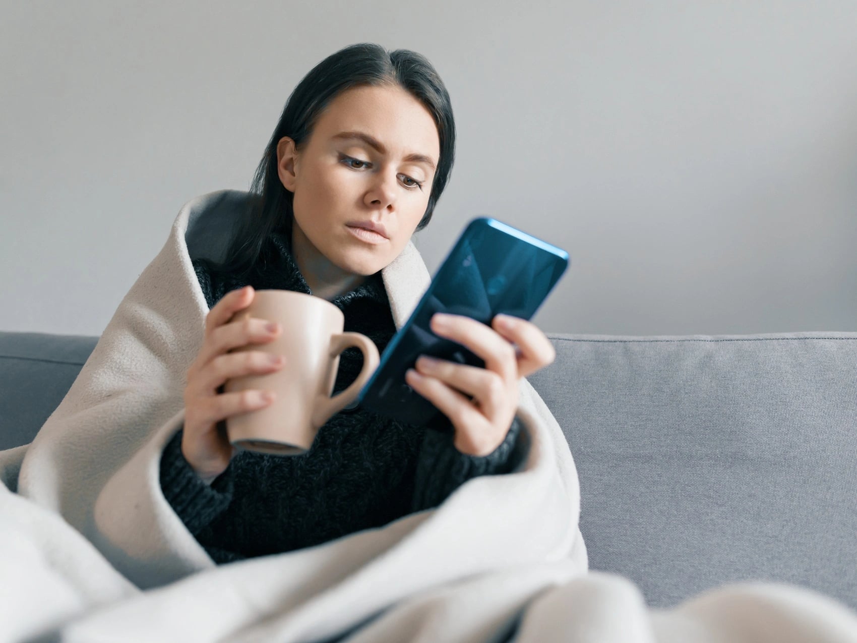Woman wrapped in a blanket on the couch holding a mug and checking a work email on her phone while home sick