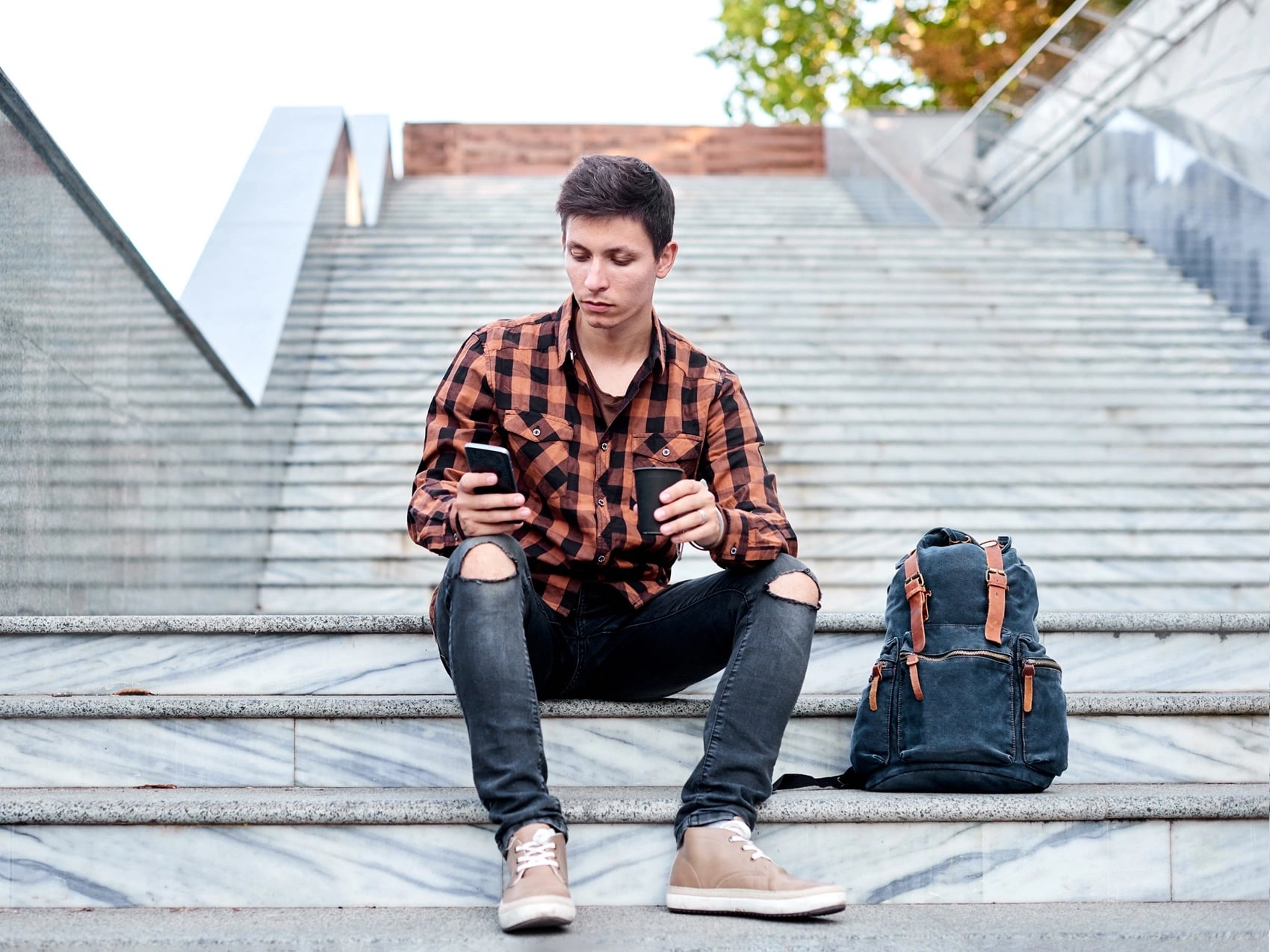 Student sitting on outdoor steps with a phone and coffee, backpack beside him