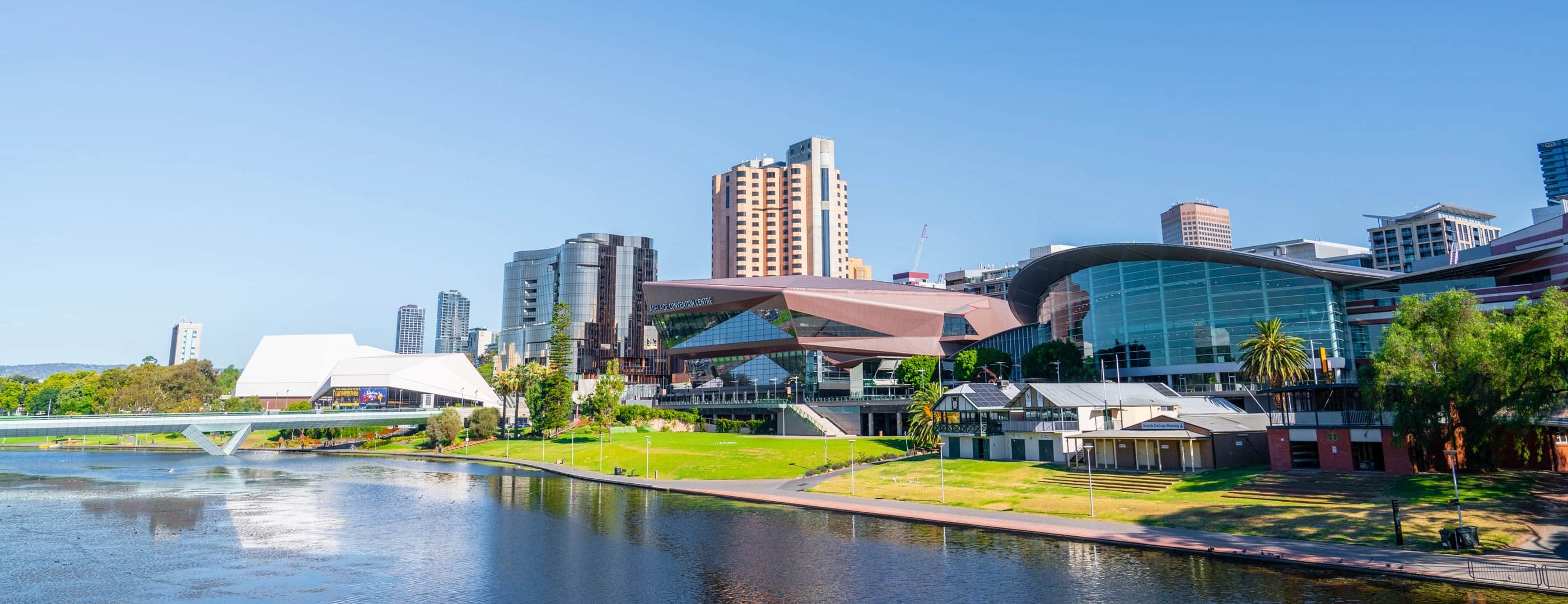 Modern city skyline beside a river, with green riverbanks and a distant bridge under a clear blue sky.