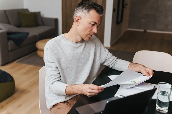 Australian man reviewing a printed medical cancellation letter at his home desk with a laptop open, checking GP requirements