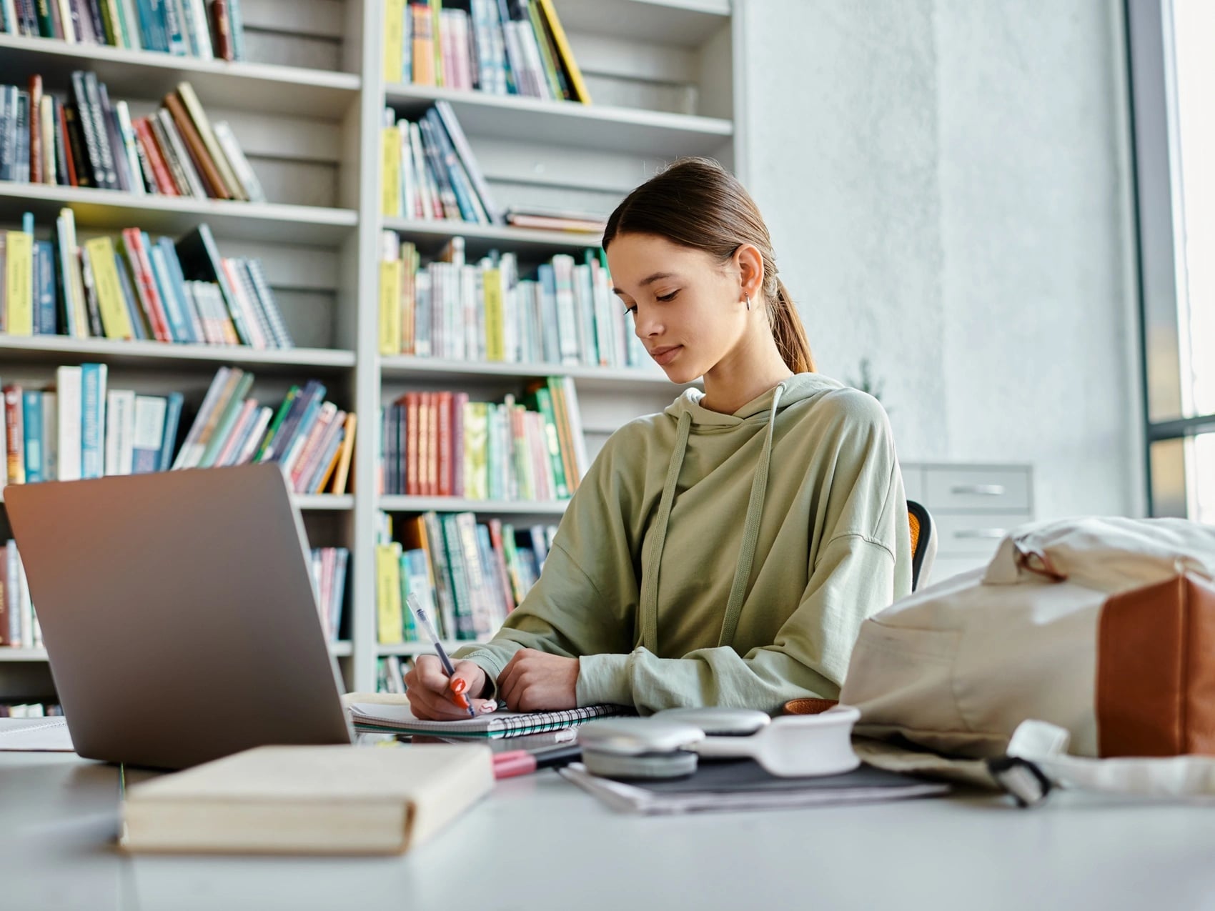 University student writing notes beside an open laptop at a desk in a quiet campus library