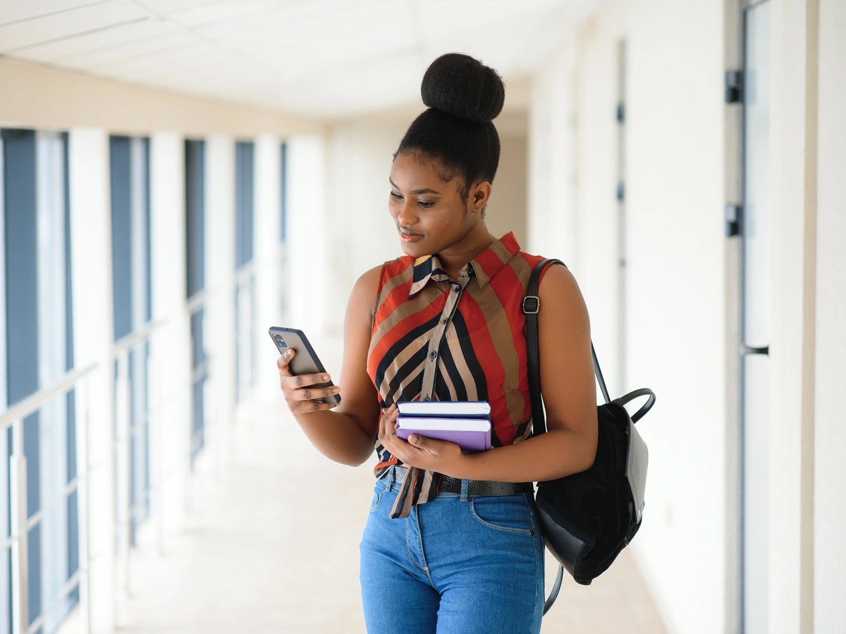 Student walking through a campus corridor, checking a phone while carrying books