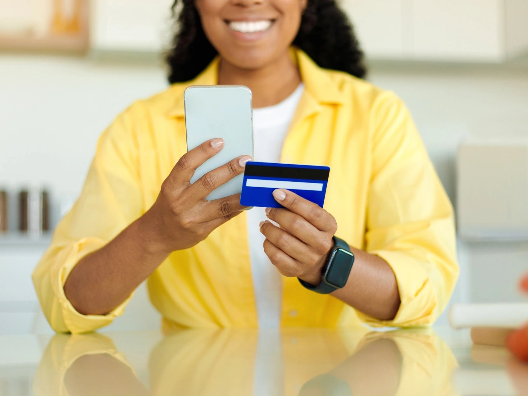 Woman in a bright kitchen holding a bank card and her phone, with no Medicare card required.