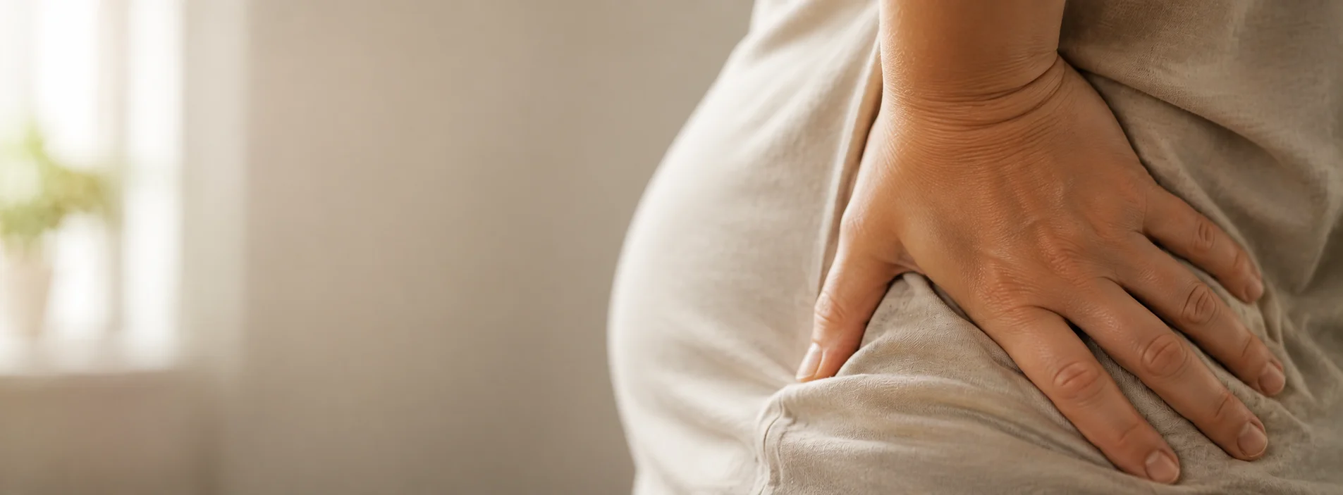 Pregnant woman pressing a hand against her lower back, showing discomfort, with soft natural light and a blurred neutral background.