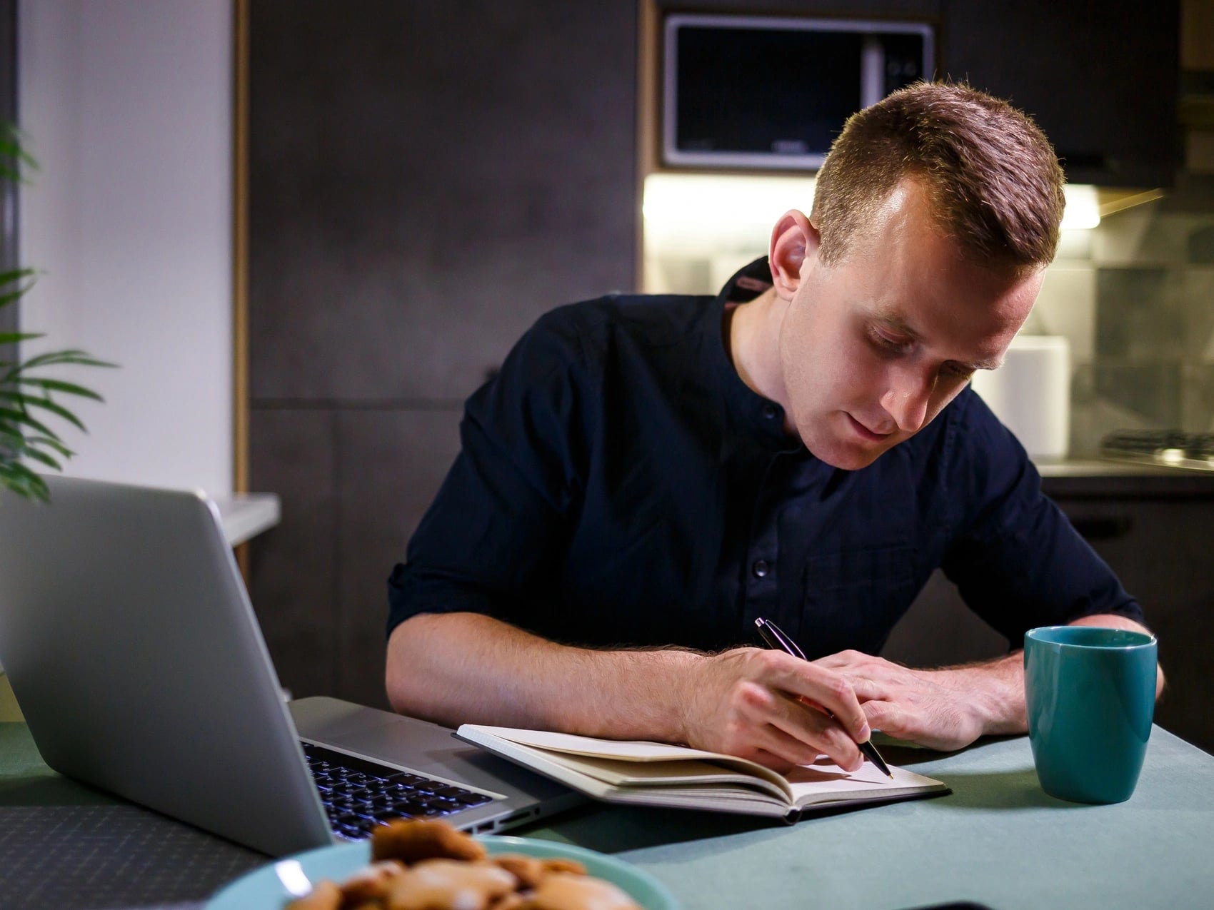 Student writing notes at a kitchen table late at night beside an open laptop