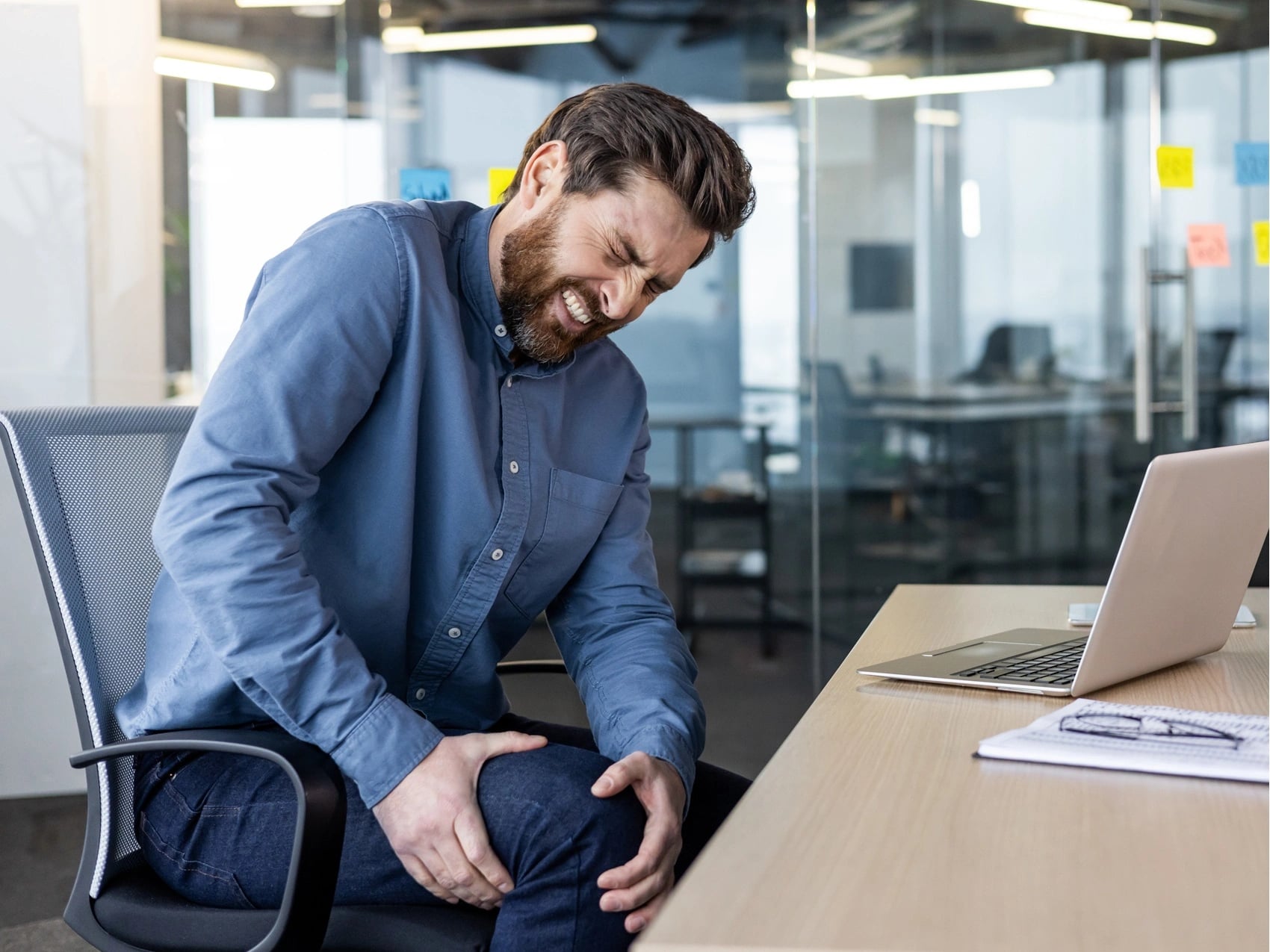 Canberra office worker holding a painful knee while seated at his desk beside a laptop